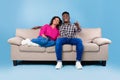 Excited young black couple with remote control sitting on couch and watching TV against blue studio background Royalty Free Stock Photo