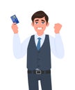 Excited young businessman showing credit, debit card. Man making or gesturing raised hand fist. Successful person in waistcoat.