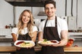 Excited young friends loving couple chefs on the kitchen holding plates with tasty food. Royalty Free Stock Photo