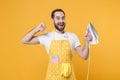 Excited young man househusband in apron hold in hands iron while doing housework isolated on yellow background studio