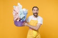 Excited young man househusband in apron throwing up basket with clean clothes while doing housework isolated on yellow