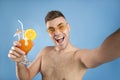 Excited young man in sunglasses holding refreshing summer cocktail and taking selfie on blue studio background Royalty Free Stock Photo
