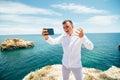 Excited young man is taking photo of himself on mobile phone. He is standing on cliff near the sea and scream Royalty Free Stock Photo