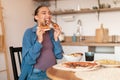 Excited young pregnant lady enjoying pizza, biting two tasty slices, sitting at table in kitchen interior, copy space Royalty Free Stock Photo