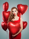 Excited young woman blowing kiss and holding balloons red heart. Beautiful girl with red lips makeup, long curly hair Royalty Free Stock Photo