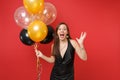 Excited young woman in little black dress celebrating spreading hands holding air balloons isolated on red background Royalty Free Stock Photo