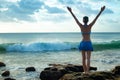 Excited young woman standing on the rock, raising arms at the beach in front of the ocean. View from back. Sunset at the beach. Royalty Free Stock Photo