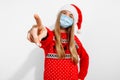 Excited young woman wearing christmas santa claus hat and medical protective mask on face pointing with index finger at camera Royalty Free Stock Photo