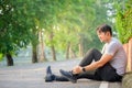 Exercise Injury Concept: A young Asian runner sits down on a park street due to pain in his ankle and grabs his sore ankle and