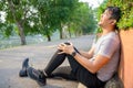 Exercise Injury Concept: A young Asian runner sits down on a park street due to pain in his knee and uses a hand to grasp the sore