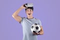 Exercising with virtual reality concept. Young athletic guy in VR headset holding soccer ball on lilac studio background