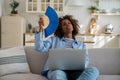 Tired mixed race woman waving fan in hand suffer from heat day while work with laptop at home couch Royalty Free Stock Photo