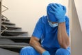 Exhausted doctor sitting on stairs indoors. Stress of health care workers during COVID-19 pandemic