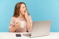 Exhausted drowsy woman sitting at workplace with laptop and yawning feeling sleepy, holding cup coffee