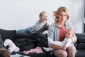 exhausted mother holding infant child and looking at camera while naughty daughter