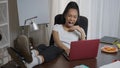 Exhausted overburdened young woman with feet on table yawning surfing internet on laptop in home office. Wide shot