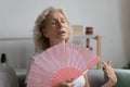 Exhausted overheated mature woman waving fan, sitting on couch