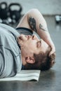 Overweight tattooed man in grey t-shirt lying on fitness mat at sports center