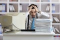 Exhausted overworked man is looking on pile of documents on desk. Paperwork concept.