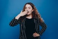 Exhausted sleepless woman has curly hair yawns covers mouth in wants to sleep awakes very early  over blue background. Royalty Free Stock Photo