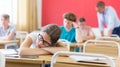 Exhausted teen girl sleeping at desk in classroom during lesson with blurred classmates