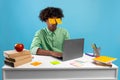 Exhausted teen guy with painted eyes on stickers sitting at desk using laptop, sleeping over blue background