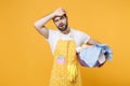 Exhausted tired young man househusband in apron hold basket with clean clothes while doing housework isolated on yellow