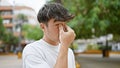 Exhausted young hispanic teenager rubbing itchy eyes amidst cool city park\'s green nature, a picture of stress in casual fashion