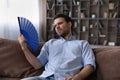 Exhausted young male recline on sofa hold fan wave himself Royalty Free Stock Photo