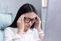 Exhaustion at work. A young Asian woman in glasses holds her head, feels pain, grimaces. Royalty Free Stock Photo