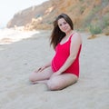 Expectant mother in red one-piece swimsuit resting on sand beach in early time Royalty Free Stock Photo