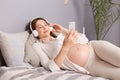 Expecting female with stunning appearance listening to music with headphones and smartphone, feeling the rhythm while revealing