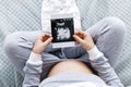 Expecting pregnant woman touching her belly while looking at ultrasound scans, sitting on cozy bed with blankets, pillows, kid Royalty Free Stock Photo