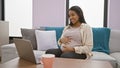 Expecting young woman relaxing and massaging belly while joyfully smiling on a video call at home