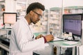 Experienced African American male pharmacist working on pc in modern pharmacy and writing working notes Royalty Free Stock Photo