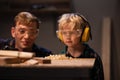 An experienced carpenter and a small boy in safety glasses blow sawdust off a work table in a carpenter`s workshop. Royalty Free Stock Photo