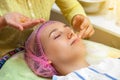 An experienced cosmetologist applies a mask of emulsion on the face of a young girl who lies on the couch during facial cleansing