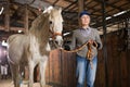 Elderly female farmer holding reins leading white horse through stable Royalty Free Stock Photo