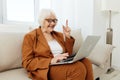 an experienced, elderly woman is sitting on a sofa in a brown suit, holding a video conference on a laptop, passing on