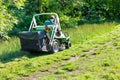 An experienced gardener mows tall grass on a tractor lawnmower while climbing the slope of a park grove, copy space