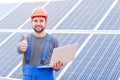 Experienced worker of the solar battery station holds a laptopand shows a thumbs-up gesture.