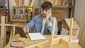 Experienced young hispanic carpenter multitasking, talking on a smartphone while using a touchpad in a busy carpentry workshop