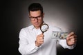 Expert authenticating 100 dollar banknote with magnifying glass against dark background, focus on hand. Fake money concept