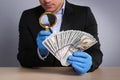 Expert authenticating 100 dollar banknotes with magnifying glass at table on light grey background, closeup. Fake money concept