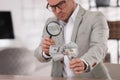 Expert authenticating 100 dollar banknotes with magnifying glass at table in office, focus on hand. Fake money