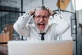 Expressive mature businessman with grey hair and beard in formal clothes is in the office against laptop