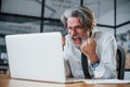 Expressive mature businessman with grey hair and beard in formal clothes is in the office against laptop