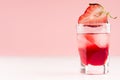 Exquisite cold red alcoholic liquor with ice cubes and strawberry slice in shot glass closeup on white wood table and pastel pink.