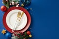 Top view featuring elegant plates, golden flatware, wine glass, ornaments, candle, confetti, frosty fir branches on blue backdrop