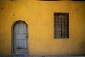 The exterior of an old yellow workplace with a locked blue door and a weathered wooden window with a rusty iron fence